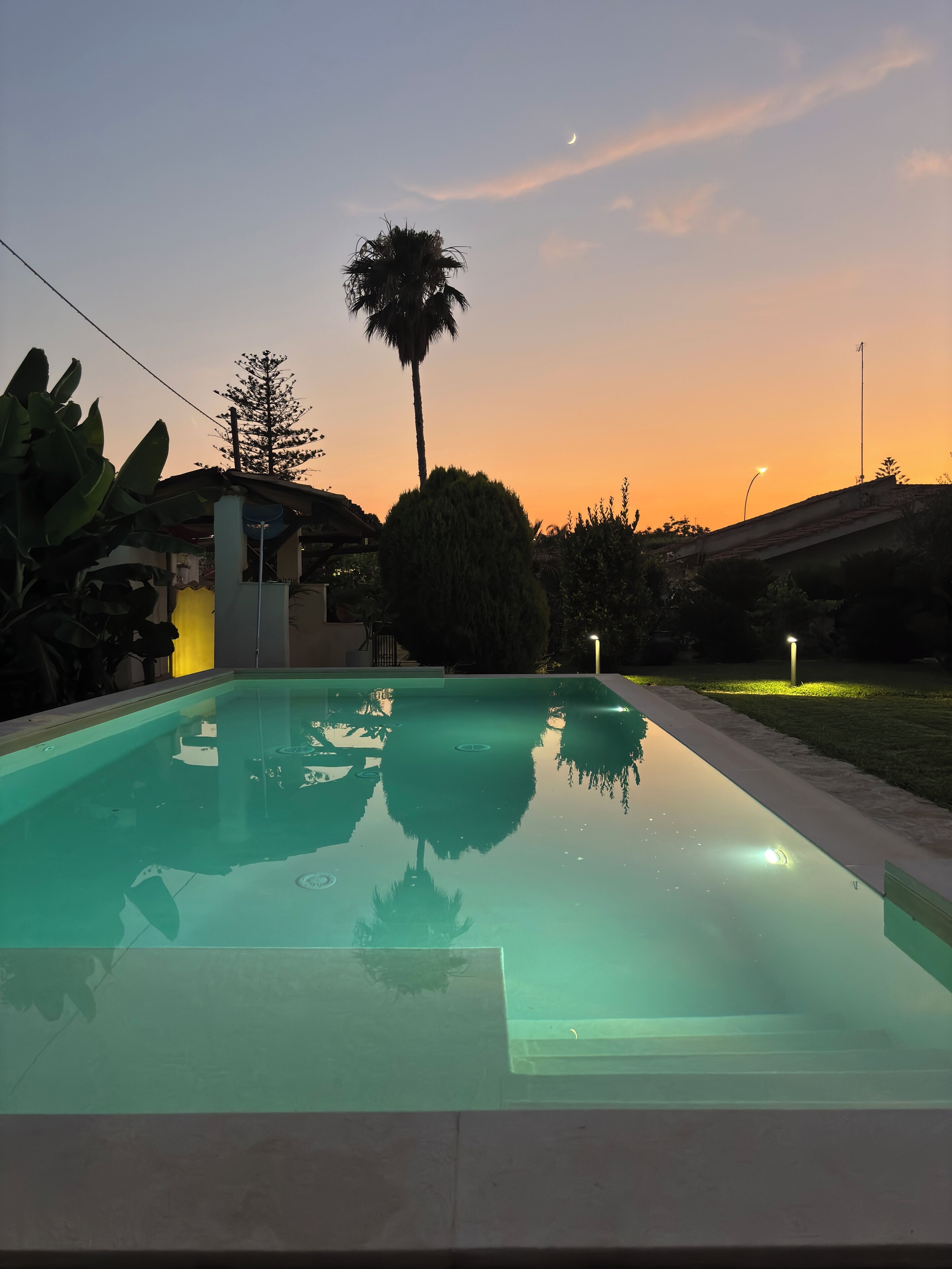 A swimming pool with clear water reflecting the surrounding trees and a sunset sky.