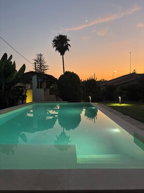 A swimming pool with clear water reflecting the surrounding trees and a sunset sky. - Villa Lucia, with an infinity pool and garden, in an oasis of peace (Siracusa)