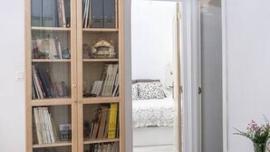 A room with a wooden bookshelf filled with books and decorative items, a white bed with a patterned bedspread, and a white door leading to another room.