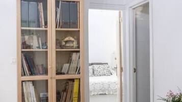 A room with a wooden bookshelf filled with books and decorative items, a white bed with a patterned bedspread, and a white door leading to another room.