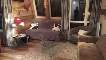 Un salon chaleureux avec un canapé gris, un fauteuil marron et une table basse en verre. On aperçoit une petite table d’appoint avec une lampe.