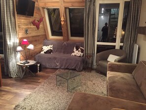 Een gezellige woonkamer met een grijze bank, een bruine fauteuil en een glazen salontafel. Een klein bijzettafeltje met een lamp is zichtbaar.