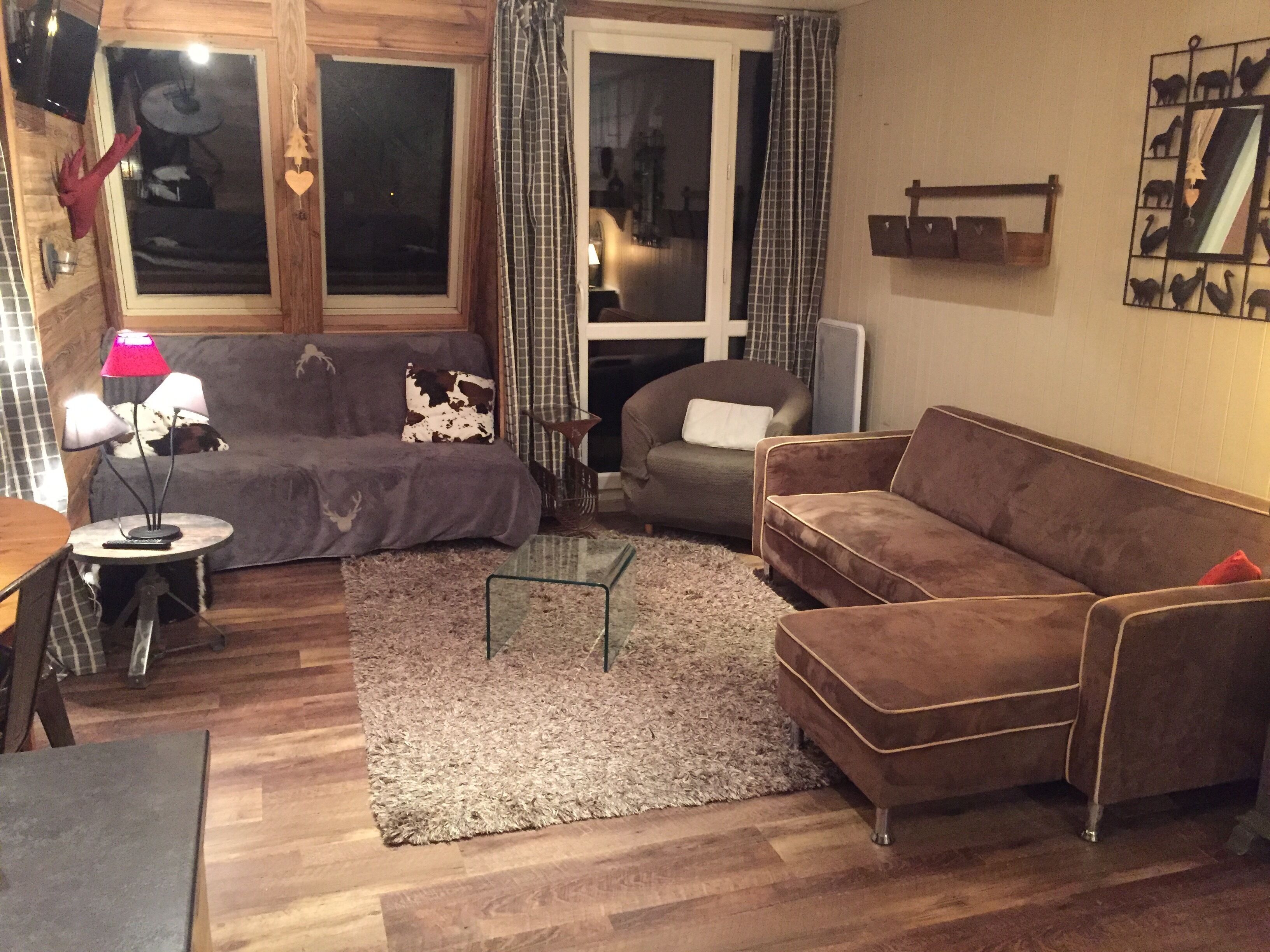 A cozy living room with a brown sofa, a glass coffee table, and a lamp. There is a window with curtains and a wall-mounted shelf with decorative items.
