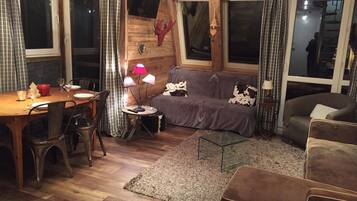 Een knus woongedeelte met een houten wand, een bank, een salontafel en een eethoek met een tafel gedekt voor twee personen.