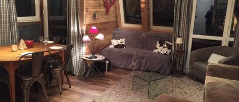 A cozy living room with a wooden wall, a sofa, a coffee table, and a dining area with a table set for two.