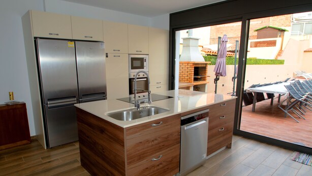 A modern kitchen with stainless steel appliances, wooden cabinetry, and a large window overlooking a patio area. - House with central pool, barbecue, wifi, a.a. and 300m from the beach. (Calella)