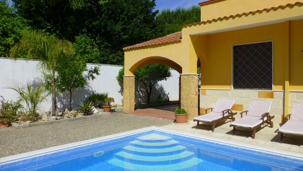 Une piscine avec des chaises longues, un patio couvert et un mur blanc agrémenté de plantes.