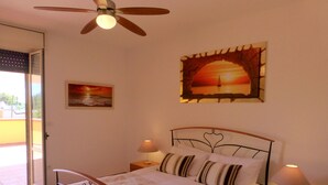 Une chambre à coucher bien rangée, avec un lit, deux tables de chevet avec des lampes, un ventilateur de plafond et un tableau représentant un coucher de soleil.