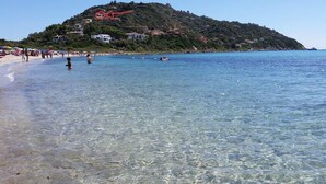 Una platja amb aigua cristal·lina, gent nedant i un turó amb edificis al fons.
