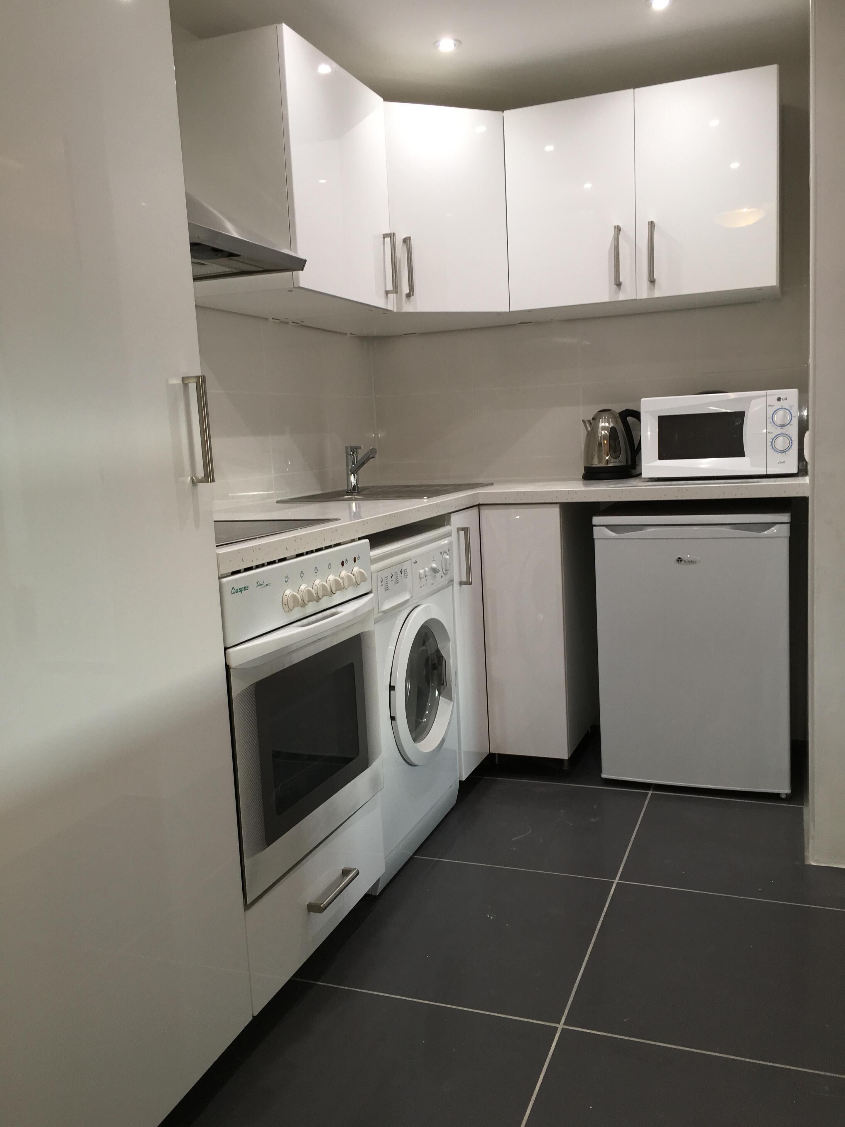 A compact kitchen with white cabinets, a built-in oven, a washing machine, a microwave, and a refrigerator.