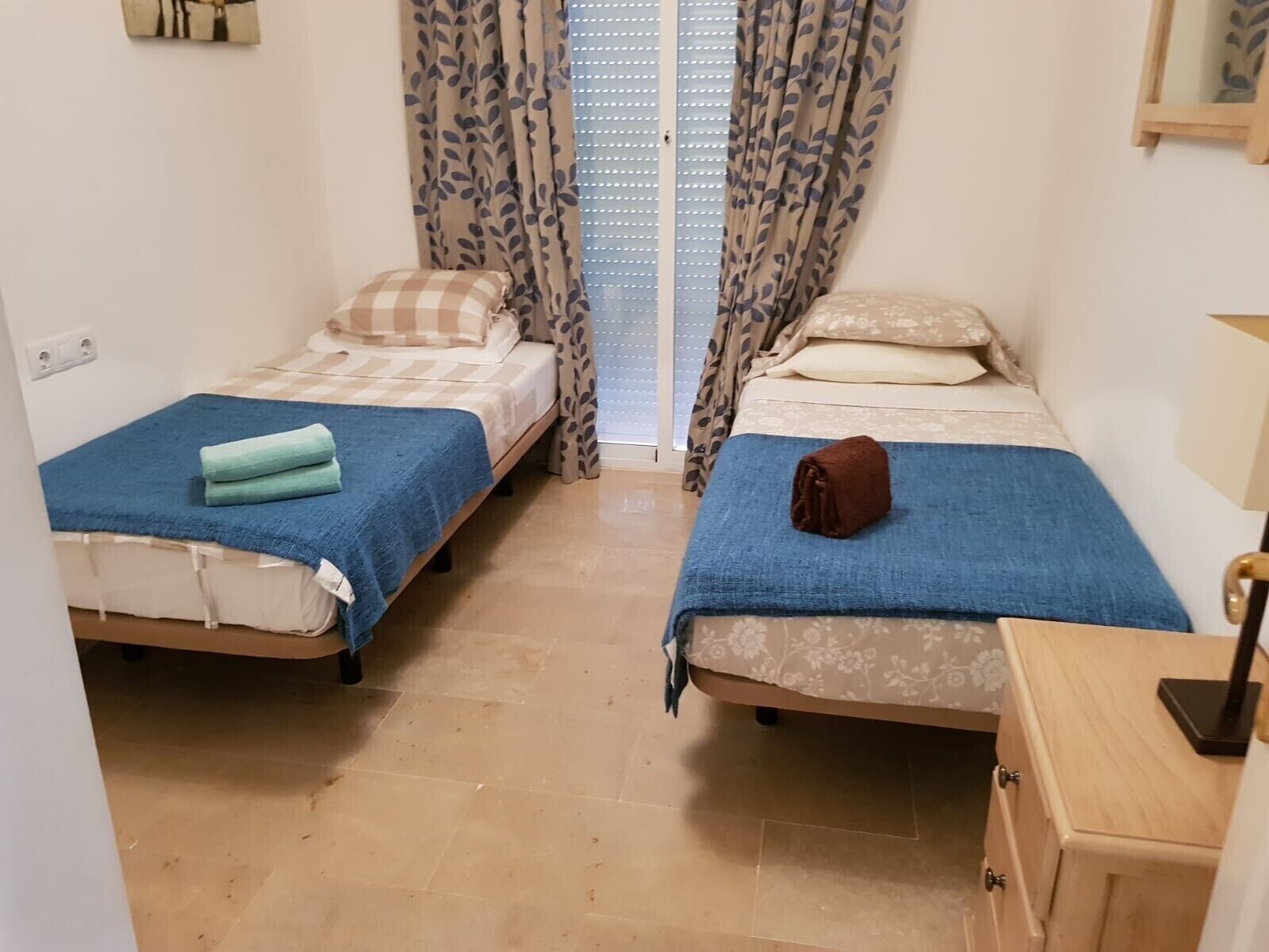 Une petite pièce, de taille modeste, avec deux lits simples, chacun recouvert d’une couverture bleue et agrémenté d’oreillers. On y trouve une armoire en bois sur laquelle est posée une lampe, ainsi qu’une fenêtre ornée de rideaux à motifs.