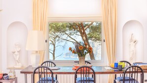 A dining area with a table set for two, a vase with flowers, and a view of trees through a window. - Villa with wonderful panoramic sea view with private pool (Capri)
