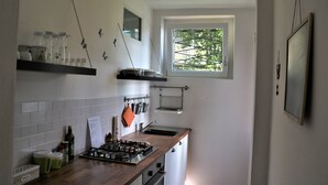 Une cuisine avec des murs recouverts de carreaux blancs, une fenêtre donnant sur des arbres, une cuisinière à gaz et divers ustensiles de cuisine.