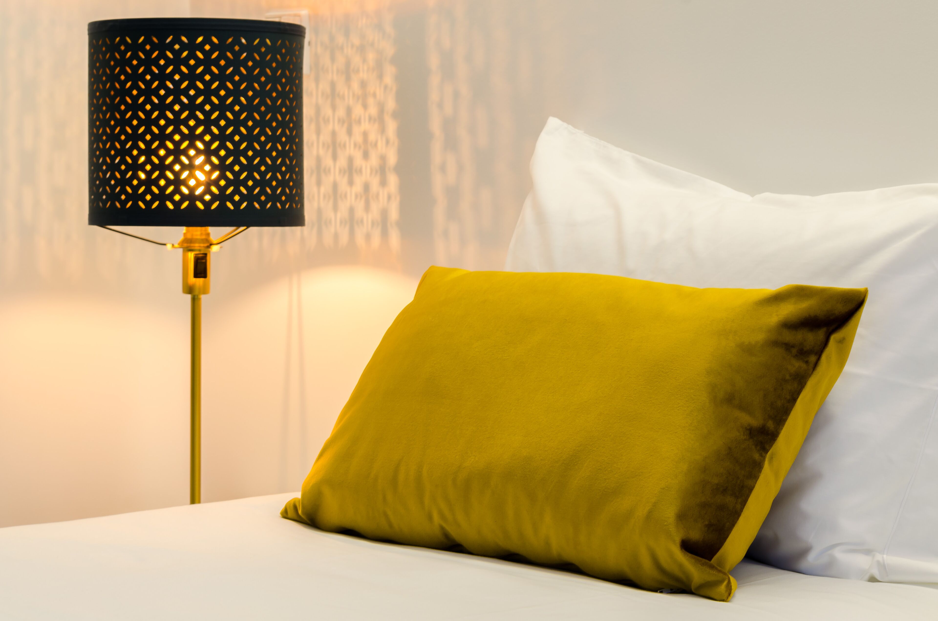 Una cama con un almohadón blanco y otro amarillo mostaza, una lámpara negra con diseño perforado, y una pared lisa de fondo.