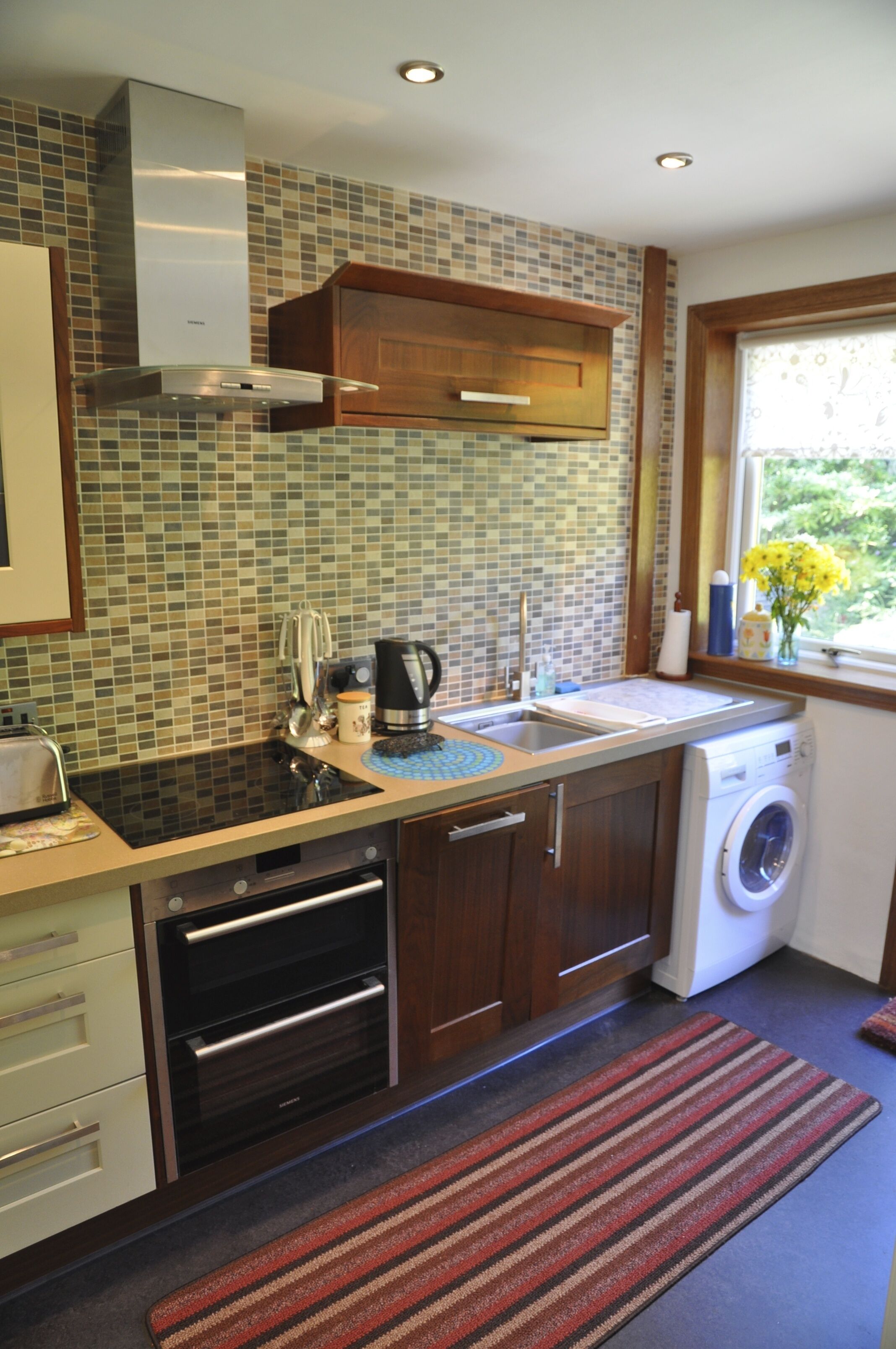 A modern kitchen with a built-in oven, a washing machine, and a window with a view of greenery.