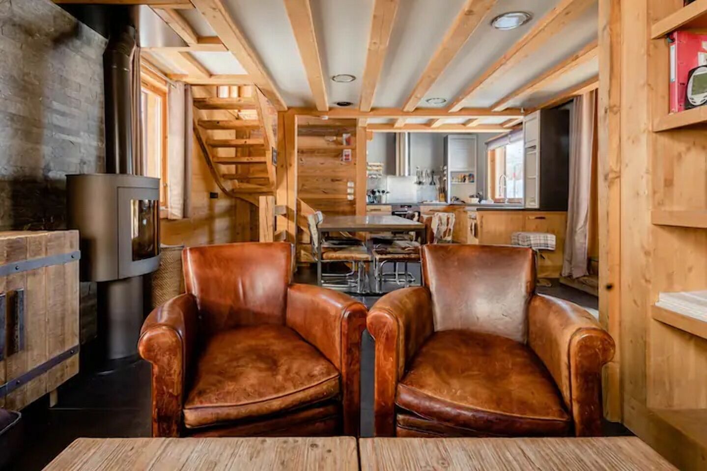 A cozy cabin interior with wooden walls, a fireplace, leather chairs, a dining table, and a kitchen area.