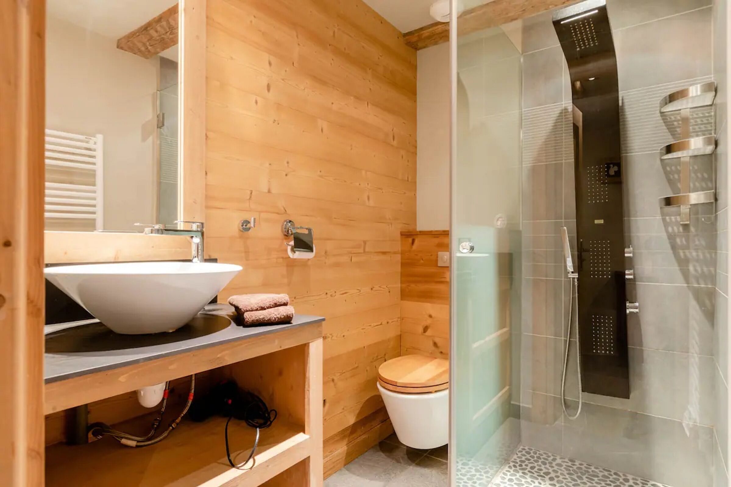 Baño de cabaña de madera con lavamanos blanco, inodoro de madera y ducha con cerramiento de vidrio.