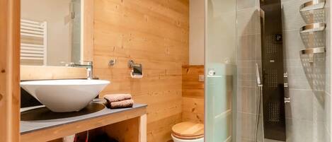 Baño de cabaña de madera con lavamanos blanco, inodoro de madera y ducha con cerramiento de vidrio.