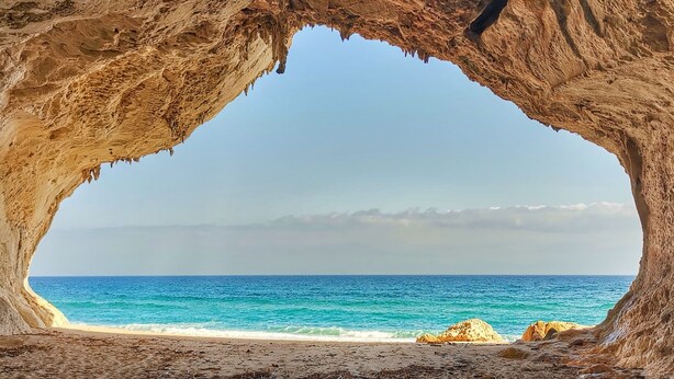 Boca de cueva que da a una playa con aguas azules cristalinas y formaciones rocosas a lo lejos.
