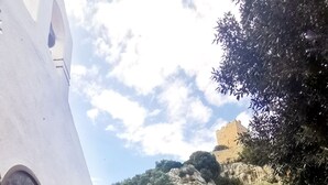 Un edificio blanco con una pared de piedra y vistas a una colina con vegetación y una estructura similar a un castillo.