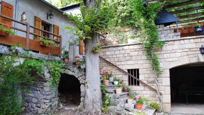 Un edificio in pietra con una scala rustica, un albero con piante rampicanti e vasi di fiori.