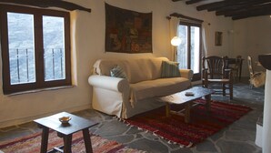 Un salon confortable avec un canapé, une table basse et un tapis. Il y a des fenêtres avec des rideaux, un tableau au mur et un coin repas avec des chaises.