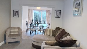 A living room with a sofa, armchair, and dining table. There are two windows with curtains, and a framed picture on the wall.