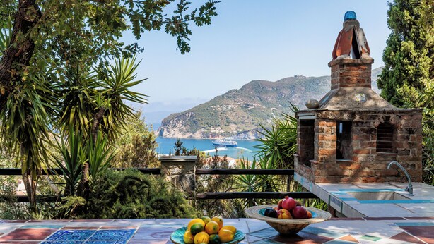 Eine Terrasse mit Steinofen, einem mit Fliesen belegten Tisch mit Obst und Blick auf Meer und Berge.