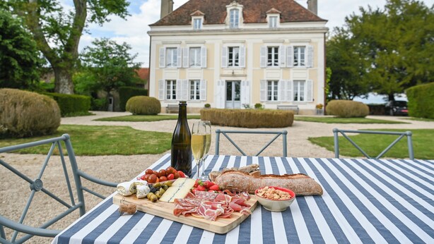 A table set with a charcuterie board, a bottle of wine, and a view of a large house in the background. - Elegant 7-Bedroom Château in Burgundy Wine Country Near Chablis & Auxerre (Hauterive)