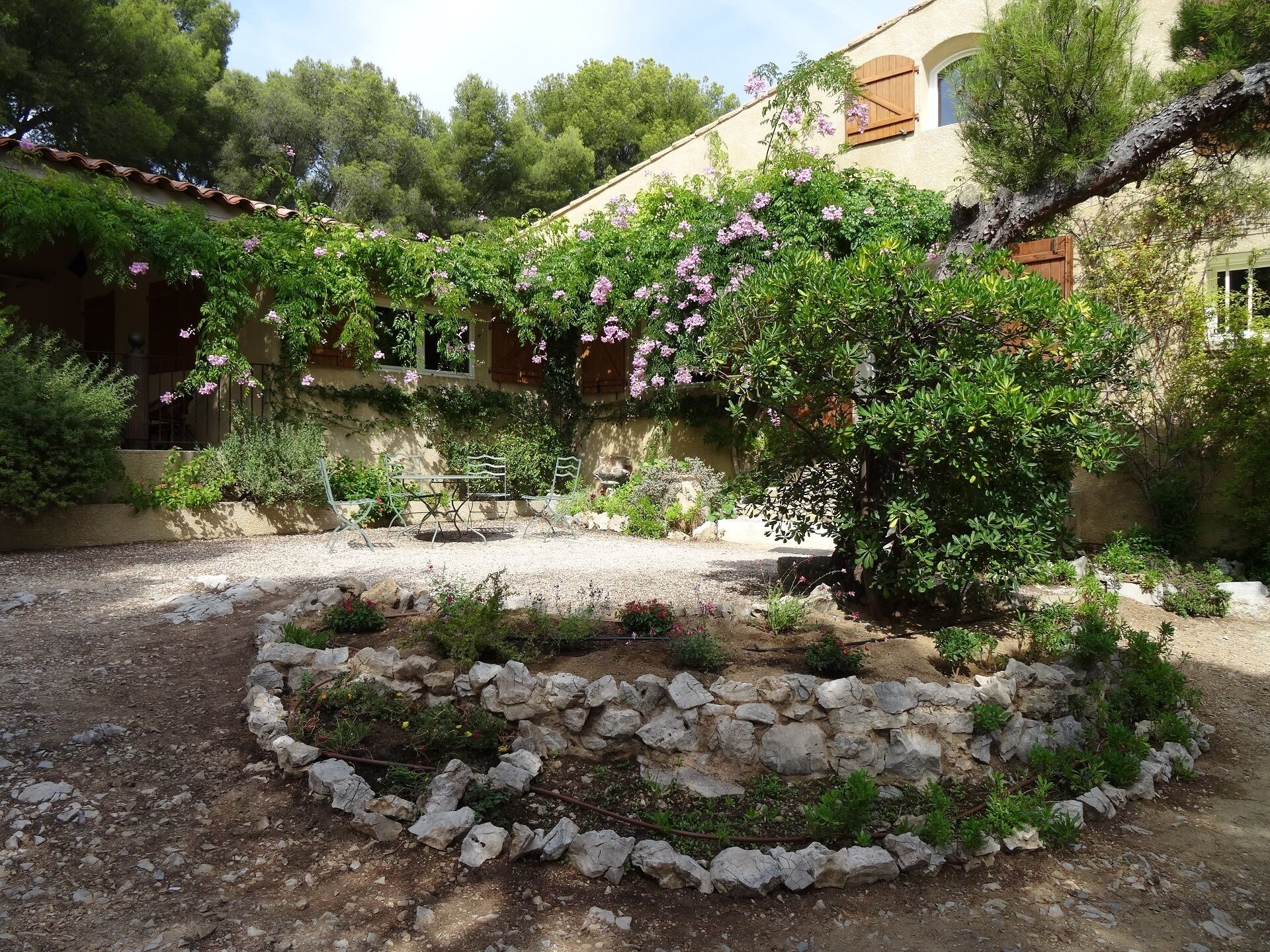 A rustic courtyard with a stone garden bed, a pergola with flowering vines, and a building with a tiled roof and wooden shutters.