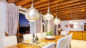 Un espace repas avec une table en bois, des chaises blanches, des luminaires décoratifs suspendus et une vue sur l’extérieur.