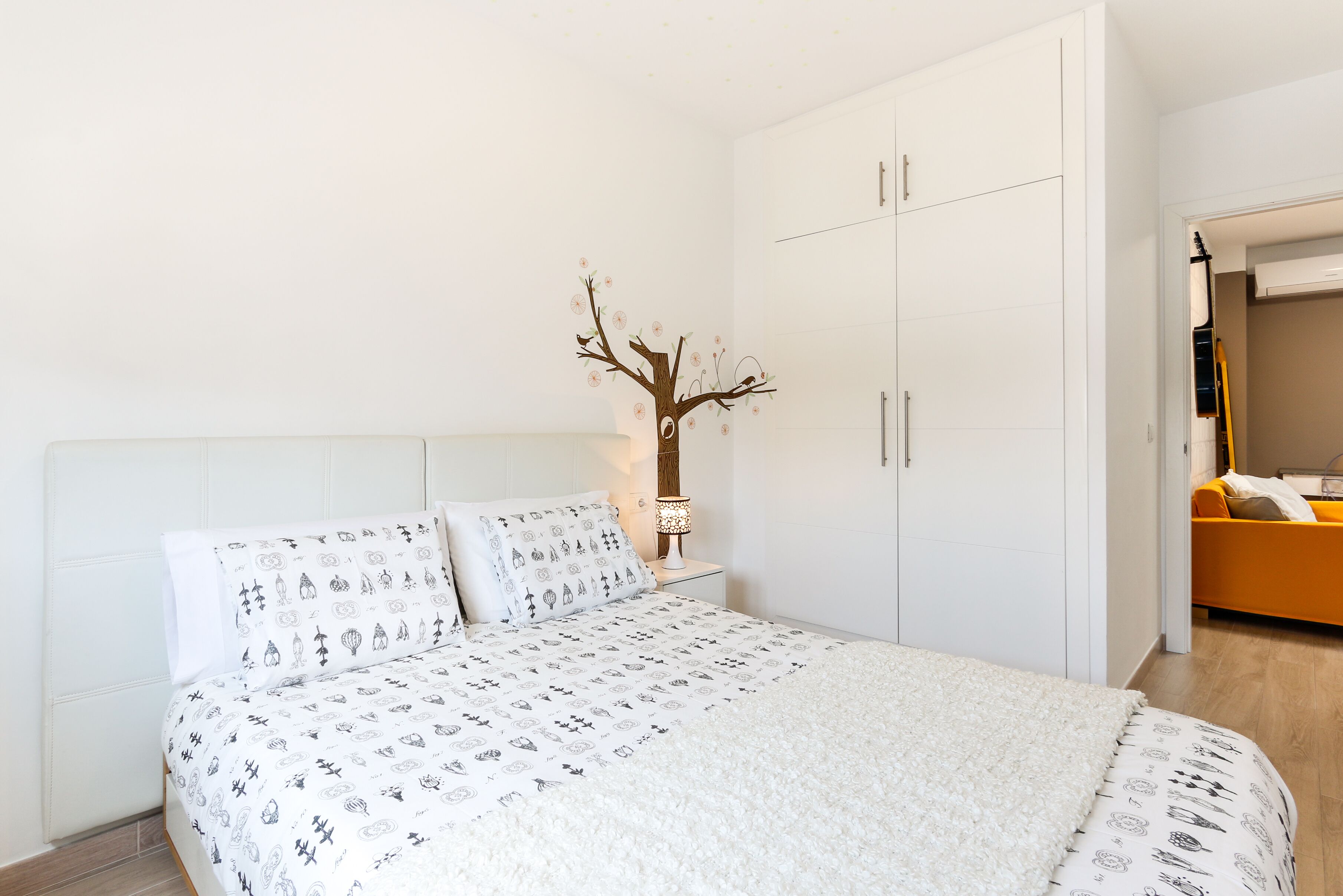 A bedroom with a bed, white headboard, a nightstand with a lamp, a wardrobe, and a small decorative tree on the wall.