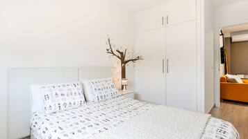 A bedroom with a bed, white headboard, a nightstand with a lamp, a wardrobe, and a small decorative tree on the wall.