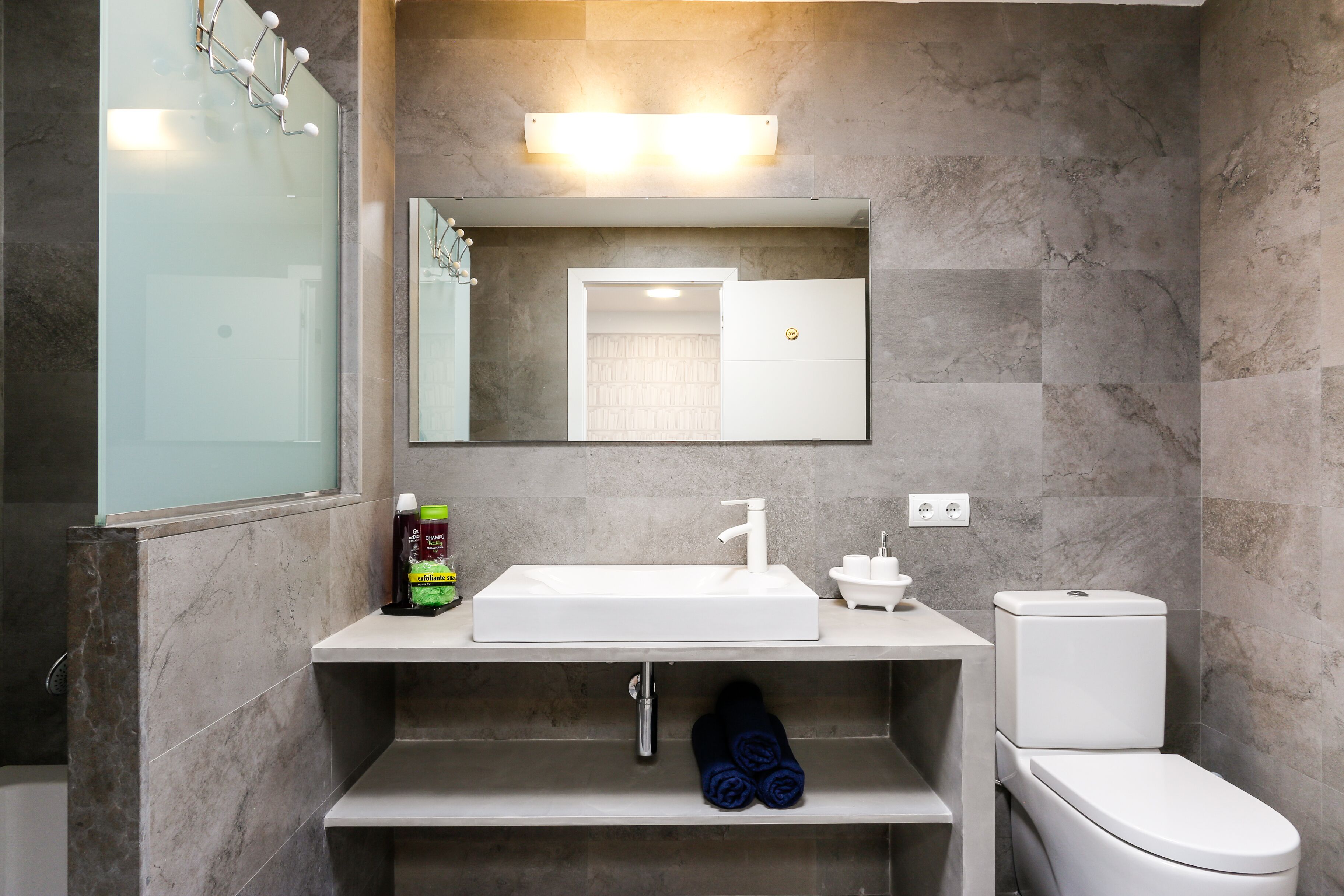 A modern bathroom with a white sink, mirror, and toilet. There is a shelf under the sink with a bottle and towels.
