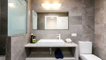 A modern bathroom with a white sink, mirror, and toilet. There is a shelf under the sink with a bottle and towels.
