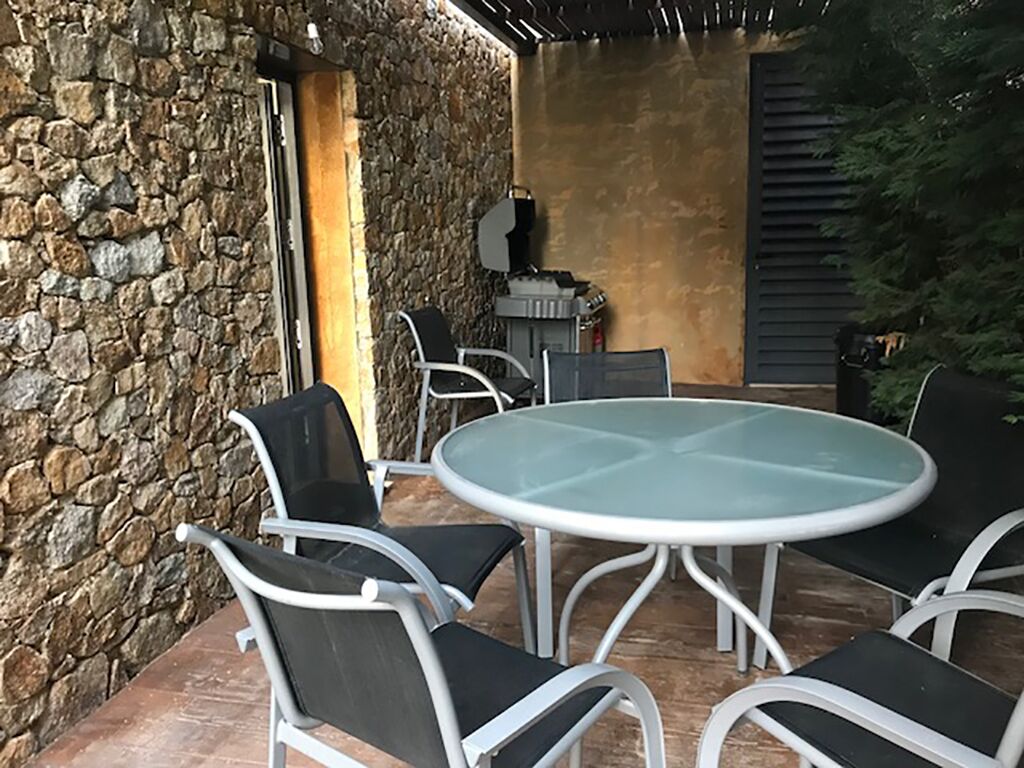 Een terras buiten met een ronde glazen tafel, vier stoelen, stenen muren en een barbecuehoek.
