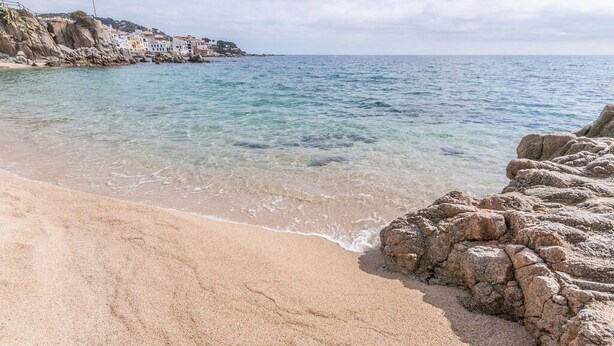 Una playa con agua cristalina, formaciones rocosas y edificios al fondo.