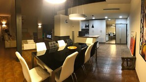 Un comedor moderno con una mesa negra y sillas blancas, una cocina al fondo y un área de estar con un sofá y televisor.