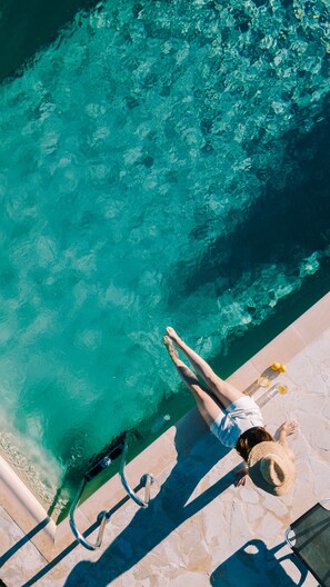 Une personne se détend au bord d’une piscine à l’eau claire, sur une terrasse en pierre, assise sur une chaise longue.