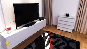 Ein modernes Wohnzimmer mit einem Flachbildfernseher auf einer weißen Konsole, einer kleinen Vase mit Blumen, einer weißen Kommode und einem gemusterten Teppich.