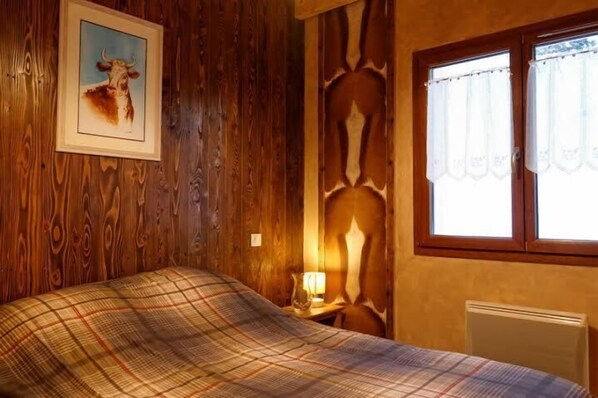 A wooden room with a bed, a framed picture of a cow, a window with curtains, and a radiator.