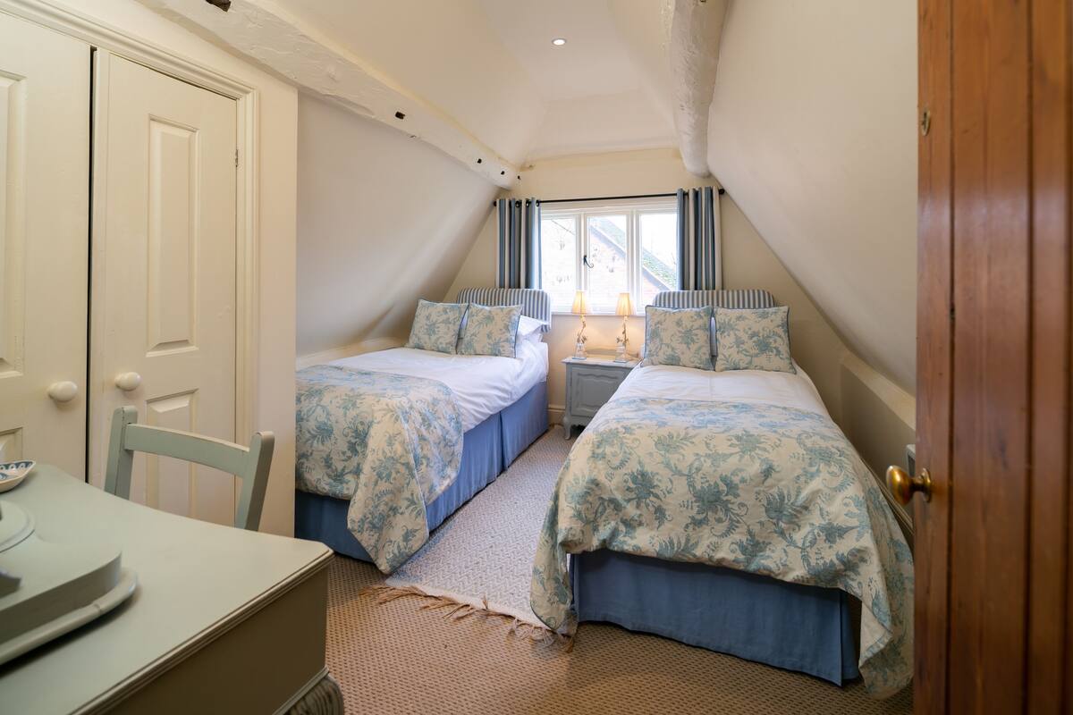 A bedroom with two beds, a window with curtains, a bedside table with lamps, and a door.