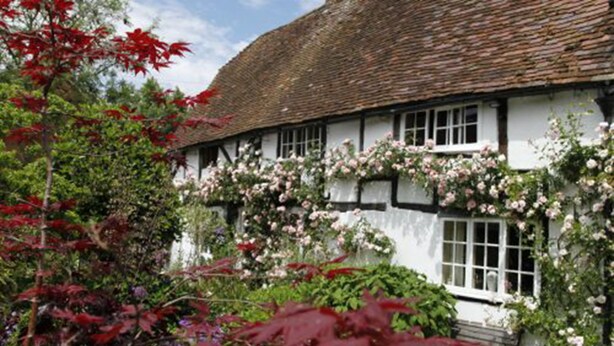 A traditional half-timbered house with a thatched roof, surrounded by lush gardens and flowering plants. - Just So Cottage (Buckingham)