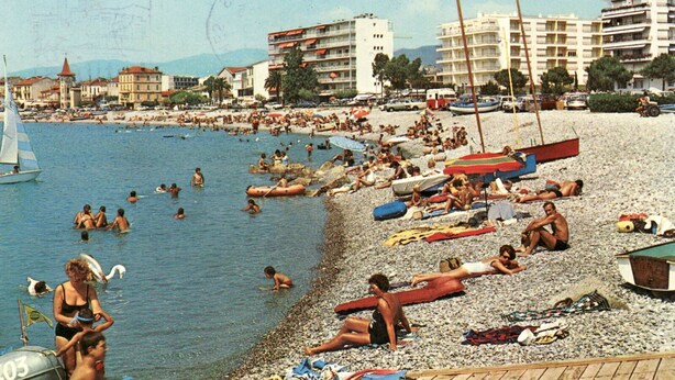 Ein überfüllter Strand mit Menschen, die sonnenbaden und schwimmen, umgeben von Gebäuden und einem Segelboot im Wasser.