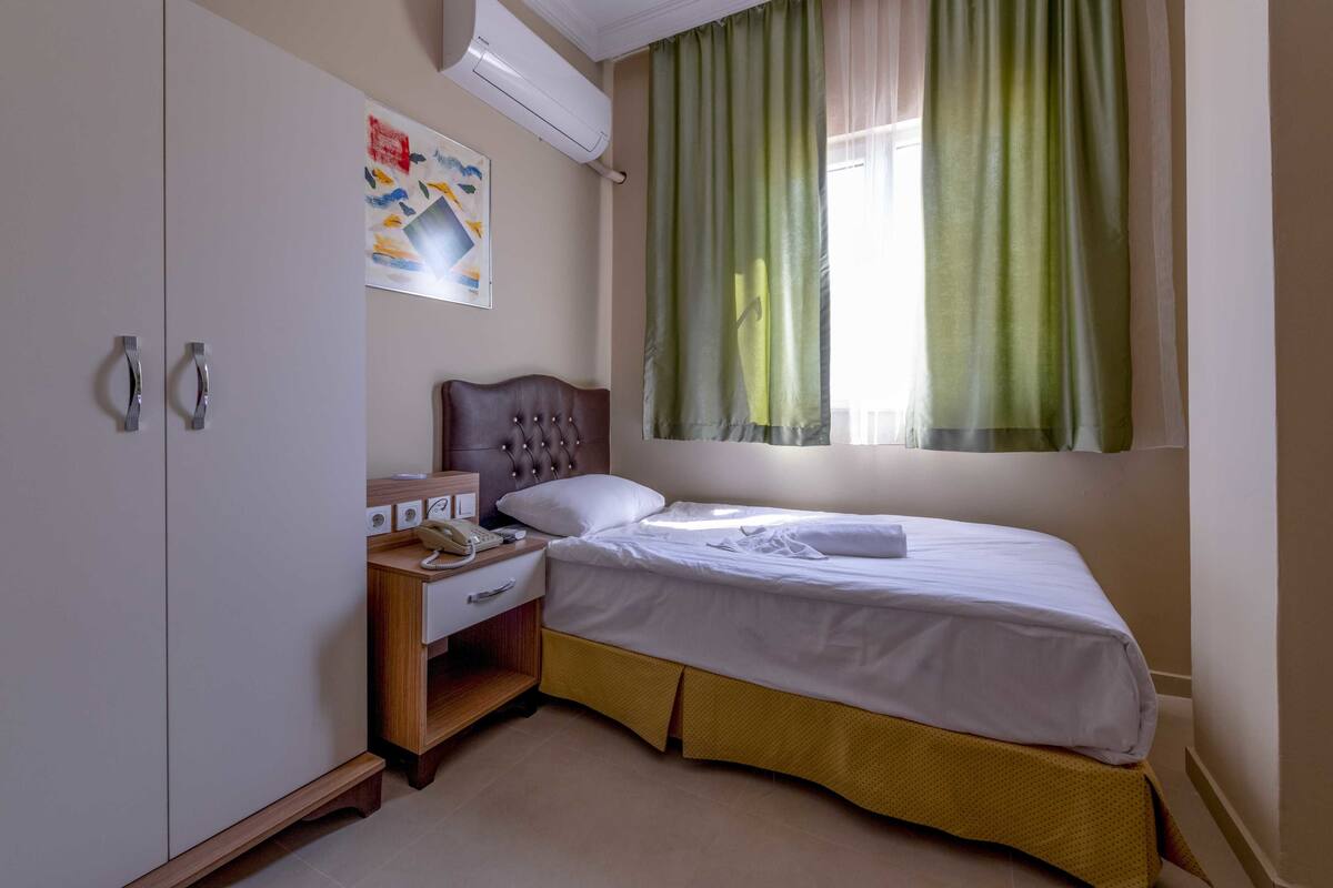 Quarto de hotel com uma cama, uma escrivaninha com telefone, um guarda-roupa e uma janela com cortinas verdes.
