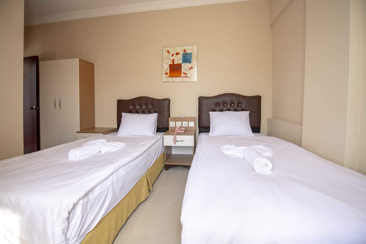 Quarto de hotel com duas camas de solteiro, um guarda-roupa de madeira, uma mesa de cabeceira com telefone e um quadro na parede.