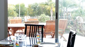 A balcony with wooden chairs and a table, overlooking a garden with trees and a fence.
