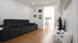 Un salon moderne avec un canapé gris foncé, une table basse blanche et un téléviseur à écran plat posé sur un meuble blanc. Des tableaux encadrés ornent le mur, et une étagère blanche est décorée de divers objets.