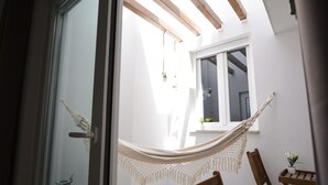 Une pièce avec un hamac, des chaises en bois, une petite table et une fenêtre ornée de plantes suspendues.