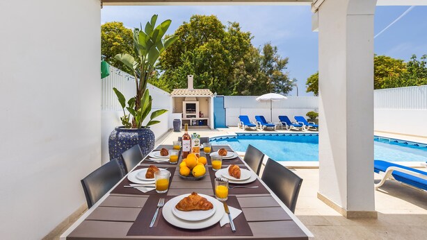 A dining table set with plates, croissants, and glasses of orange juice, overlooking a swimming pool and a garden.
